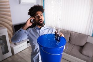 man-on-phone-while-holding-bucket-up-under-ceiling-collecting-water