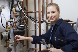 Smiling technician performing heat pump maintenance surrounded by copper pipes and control valves.